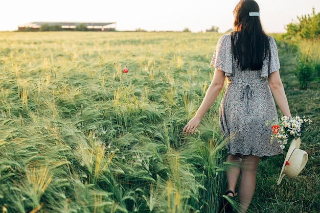 Beautiful woman with wildflowers and straw hat walking in barley field in sunset light Stylish female relaxing in evening summer countryside and gathering flowers Atmospheric tranquil moment