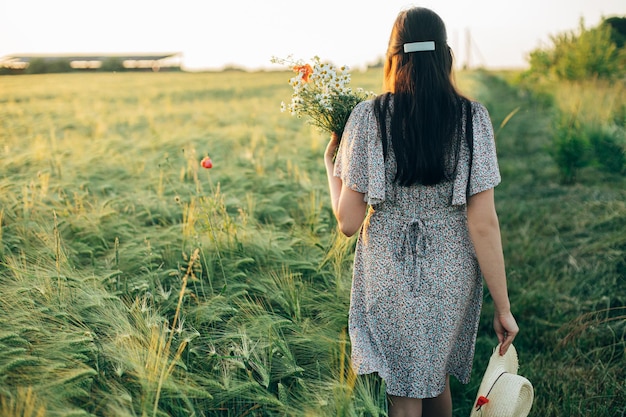 Beautiful woman with wildflowers and straw hat walking in barley field in sunset light Stylish female relaxing in evening summer countryside and gathering flowers Atmospheric tranquil moment