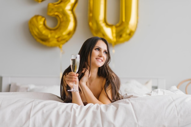 Beautiful woman woke up in bed in the morning and celebrates her thirties with a glass of champagne in a nightgown