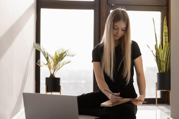 Beautiful woman working at home at the computer sitting on the floor by the window and making notes in a notebook