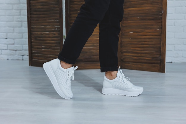 Beautiful womens shoes with legs leather white sneakers in the interior of a room or office shoes for a woman black trousers and white sneakers casual and modern style modern and trendy