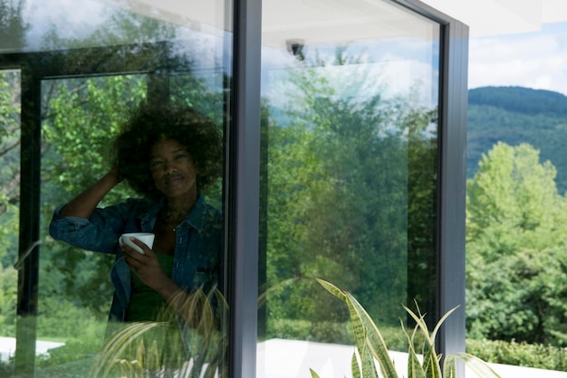 Beautiful young african american woman drinking coffee and looking through a window in her luxury home