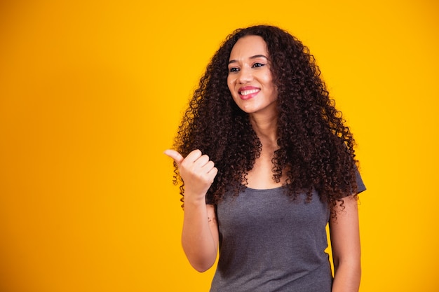 Beautiful young African American woman with curly hair making a happy thumbs-up gesture with her hand. Approval expression looking at camera showing success.