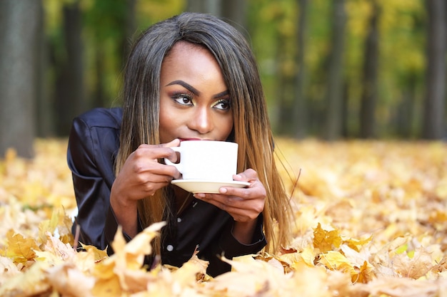 Beautiful young African woman drinking coffee from a white Cup on the of autumn leaves in the Park