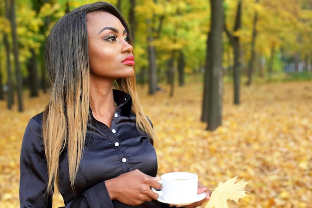 Beautiful young African woman drinking coffee from a white Cup on the background of autumn leaves in the Park