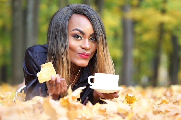 Beautiful young African woman drinking coffee from a white Cup on the surface of autumn leaves in the Park