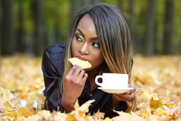 Beautiful young African woman drinking coffee from a white Cup on the surface of autumn leaves in the Park