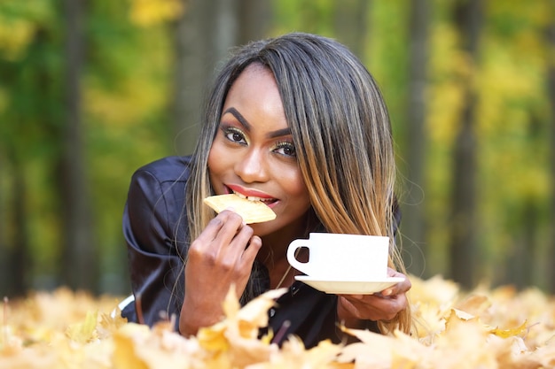 Beautiful young African woman eating cookies and drinking coffee from white Cup on autumn leaves  in Park