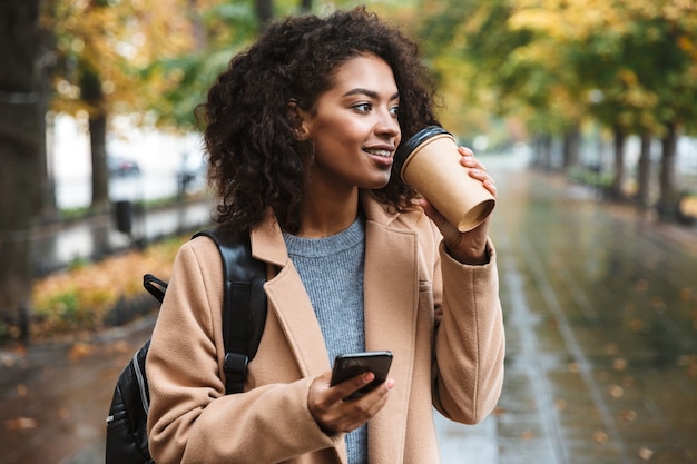 Beautiful young african woman wearing coat walking outdoors at the park, carrying backpack, using mobile phone, drinking coffee