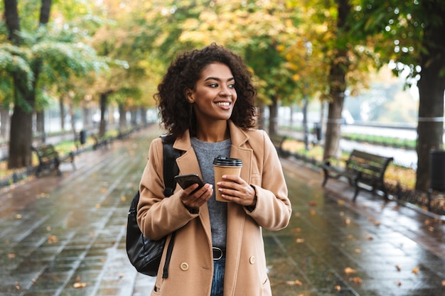 Beautiful young african woman wearing coat walking outdoors at the park, carrying backpack, using mobile phone, drinking coffee