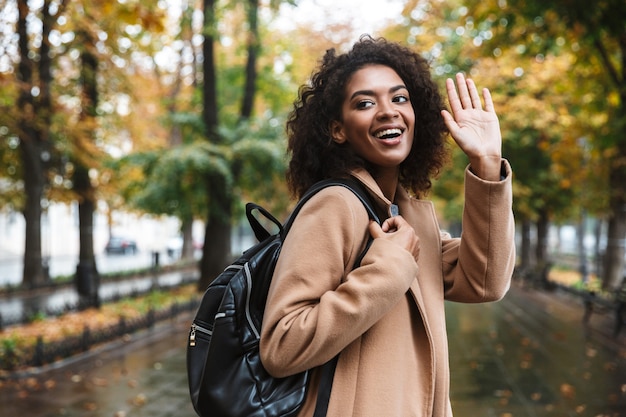 Beautiful young african woman wearing coat walking outdoors at the park, carrying backpack