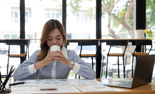 Beautiful young Asian businesswoman smiling holding a coffee mug and laptop working at the office