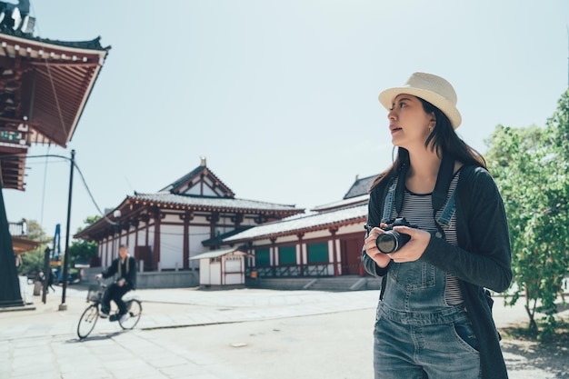 Beautiful young asian lady tourist at traditional japanese temple shitennoji in osaka japan. girl standing under blue sky hold camera photographing while traveling. male in suit riding bike bicycle.