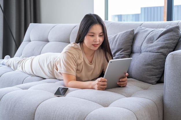 beautiful young asian woman in casual cozy clothes using tablet sitting on couch at modern apartment