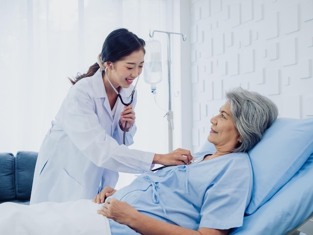 A beautiful young Asian woman doctor in white suit smiled while using stethoscope to examine listen to heartbeat of elderly old female patient in blue dress who was lying on bed in hospital room