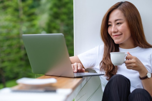 A beautiful young asian woman drinking coffee while using and working on laptop computer in the outdoors