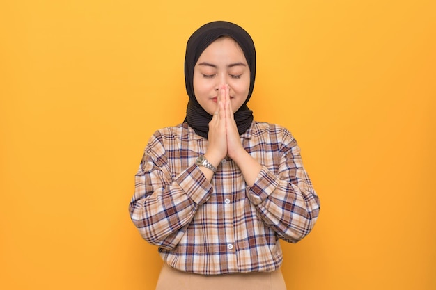 Beautiful young Asian woman in plaid shirt makes a praying gesture with closed eyes isolated on yellow background