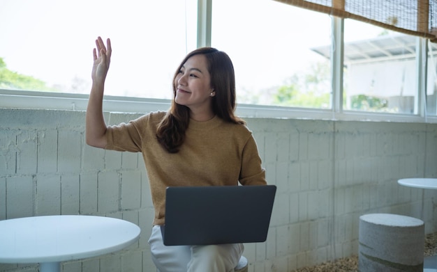 A beautiful young asian woman raising hand to say hi to other people while using laptop computer