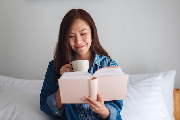 A beautiful young asian woman reading book and drinking hot coffee in a white cozy bed at home