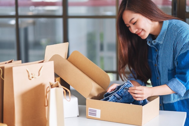 A beautiful young asian woman receiving and opening a postal parcel box of clothing at home for delivery and online shopping concept