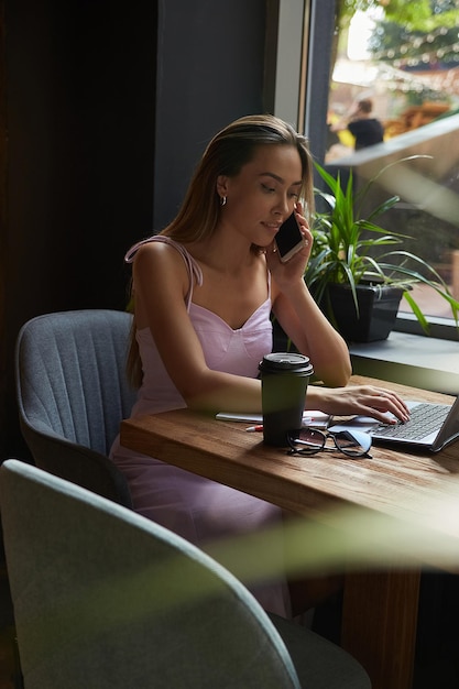 Beautiful young asian woman sitting at coffee shop table surrounded by plants working on laptop