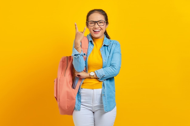 Beautiful young Asian woman student in denim outfit with backpack shouting with crazy expression doing rock symbol with hands up isolated on yellow background  Education in university college concept