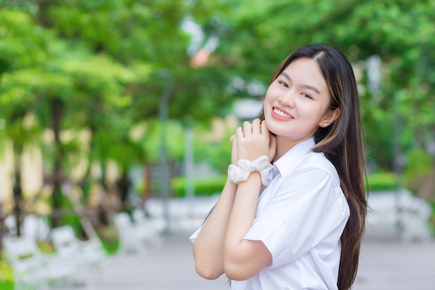 Beautiful young Asian woman student who has blond long hair smiles and looks at to camera while holding hands and standing in university with outdoor tree background.