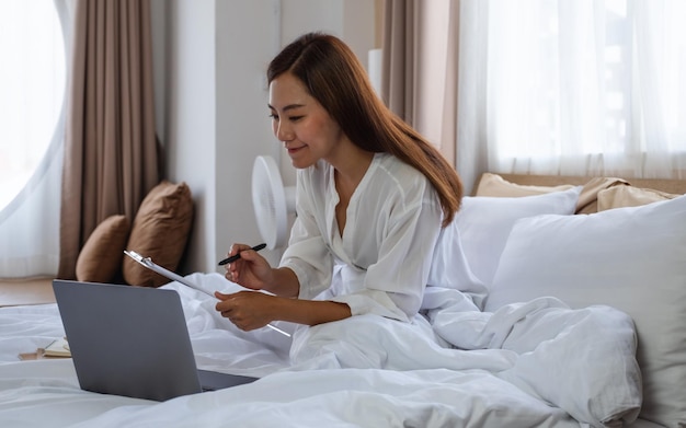 A beautiful young asian woman using laptop computer and working on paperwork while sitting on bed at home