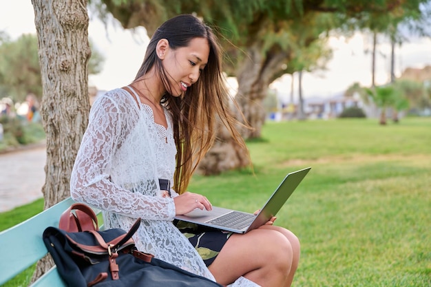 Beautiful young Asian woman using laptop sitting on an outdoor bench