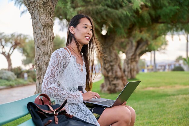 Beautiful young Asian woman using laptop sitting on an outdoor bench