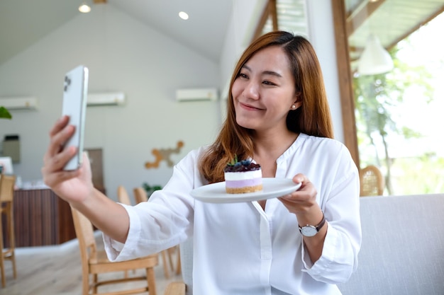 A beautiful young asian woman using mobile phone to take a selfie with a cake before eat in cafe