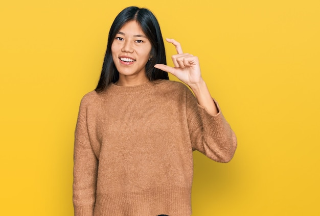 Beautiful young asian woman wearing casual winter sweater smiling and confident gesturing with hand doing small size sign with fingers looking and the camera measure concept