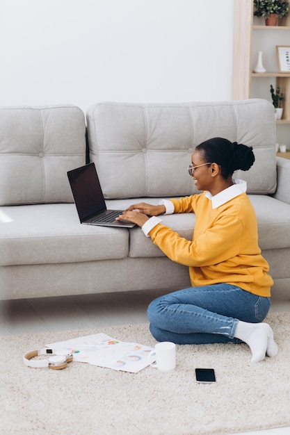 Beautiful young Black female university student sitting on the floor, drinking coffee and doing project on laptop