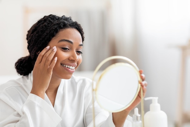 Beautiful young black lady looking in mirror and applying under eye cream