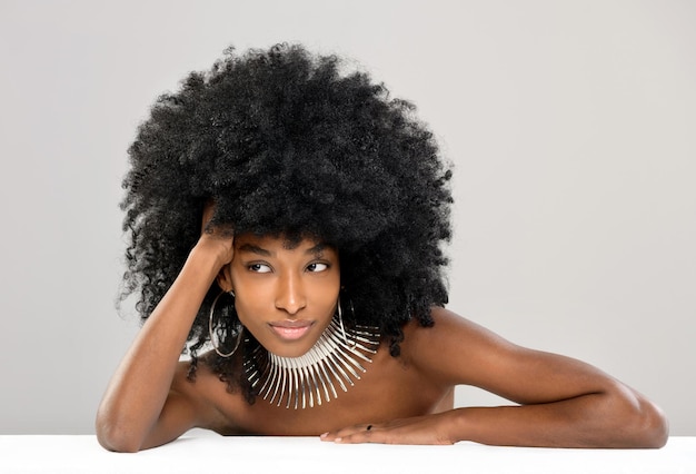 Beautiful young Black woman modelling a modern silver choker or necklace with hoop earrings leaning over a table with her bare shoulders looking aside with a quiet smile