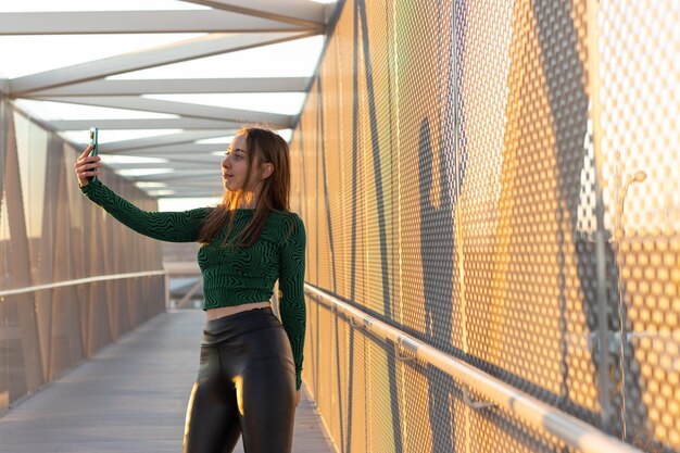 A beautiful and young blonde and Caucasian woman stands on a bridge to take a selfie on her mobile phone in a sunny day during the sunset