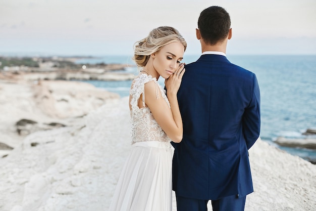 Beautiful young blonde model girl in fashionable white lace dress lean on the handsome man in the stylish blue suit and posing on the white rock at the coast of the Adriatic sea