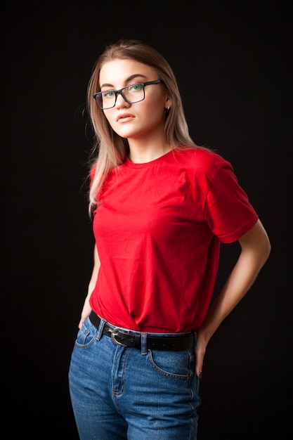 Beautiful young blonde woman in black glasses, red t-shirt and jeans posing