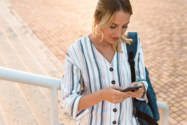 Beautiful young blonde woman carrying backpack using mobile phone while standing on steps outdoors