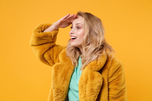 Beautiful young blonde woman girl in yellow fur coat posing isolated on orange background in studio. People lifestyle concept. Mock up copy space. Holding hand at forehead looking far away distance.