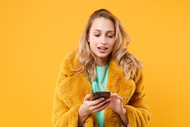 Photo beautiful young blonde woman girl in yellow fur coat posing isolated on orange background studio portrait. people emotions lifestyle concept. mock up copy space. using mobile phone typing sms message.