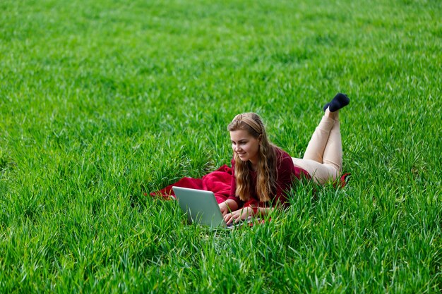 Photo beautiful young blonde woman is lying on the green grass in the park with a laptop and working. blue sky with clouds. the girl smiles and enjoys a good day. work on the nature on a sunny day.