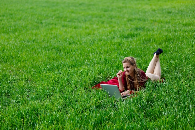 Photo beautiful young blonde woman is lying on the green grass in the park with a laptop and working. blue sky with clouds. the girl smiles and enjoys a good day. work on the nature on a sunny day.