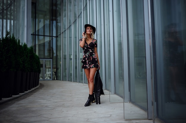 A beautiful young blonde woman in a leather black jacket, a pretty short dress and a hat walks in the Zaryadye Park, Moscow. 