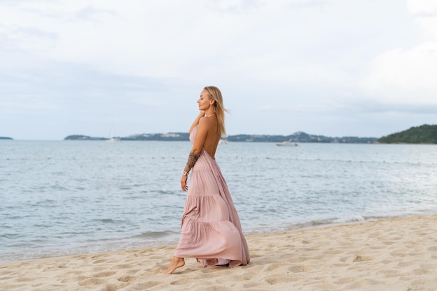 Beautiful young blonde woman in a long pink dress on the beach tender posing the wind develops the dress hair romantic free