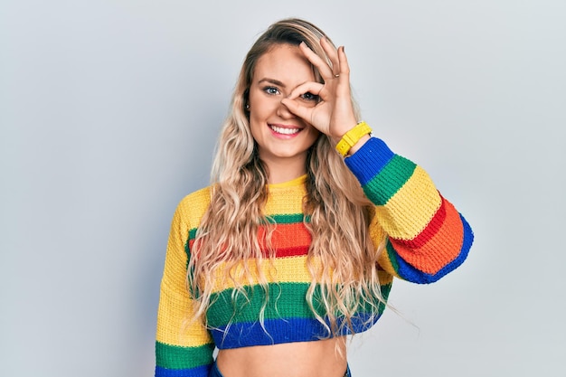 Beautiful young blonde woman wearing colored sweater doing ok gesture with hand smiling eye looking through fingers with happy face