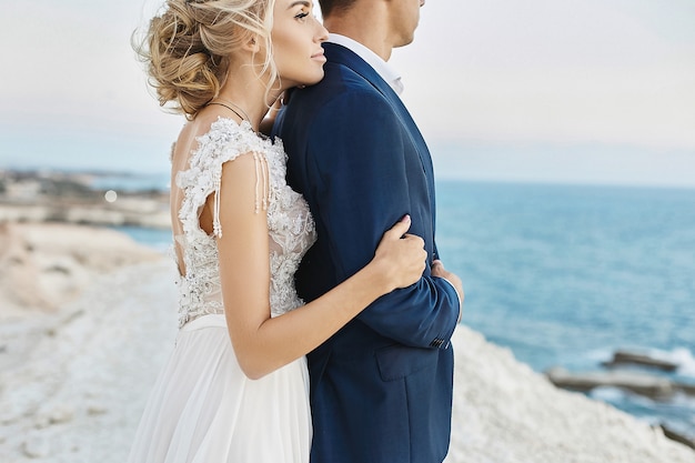 Beautiful young blonde woman in white lace dress leans on the handsome man in the stylish blue suit on the white rock at the coast of the Adriatic sea. Model girl in a wedding dress with stylish groom