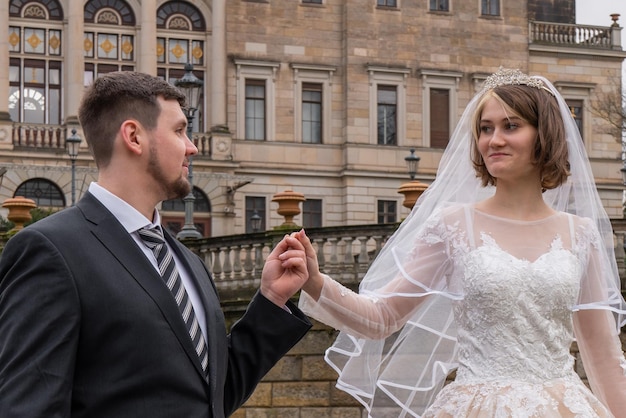 A beautiful young bride tenderly looks at the groom The groom holds her hand Walk on the background of an ancient castle buildings Love and harmony young family