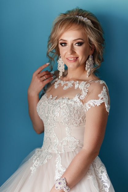 Beautiful young bride with hairstyle and with white gold tiara on her head in white long dress on blue wall background.