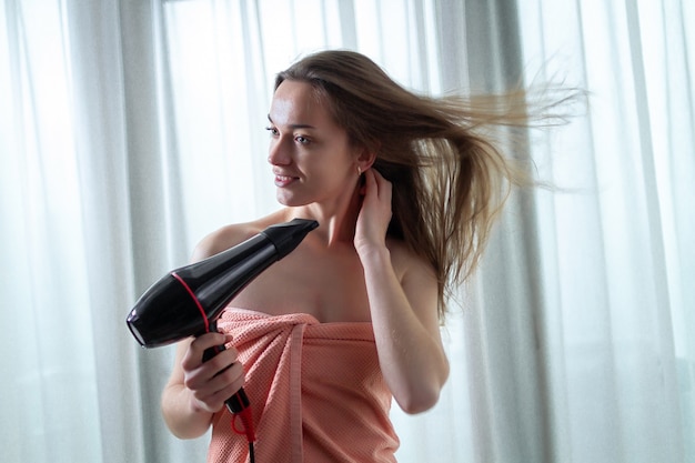 Beautiful young brown haired person in bath towel is using a hair dryer for drying her long hair after shower in room. Hair Care.